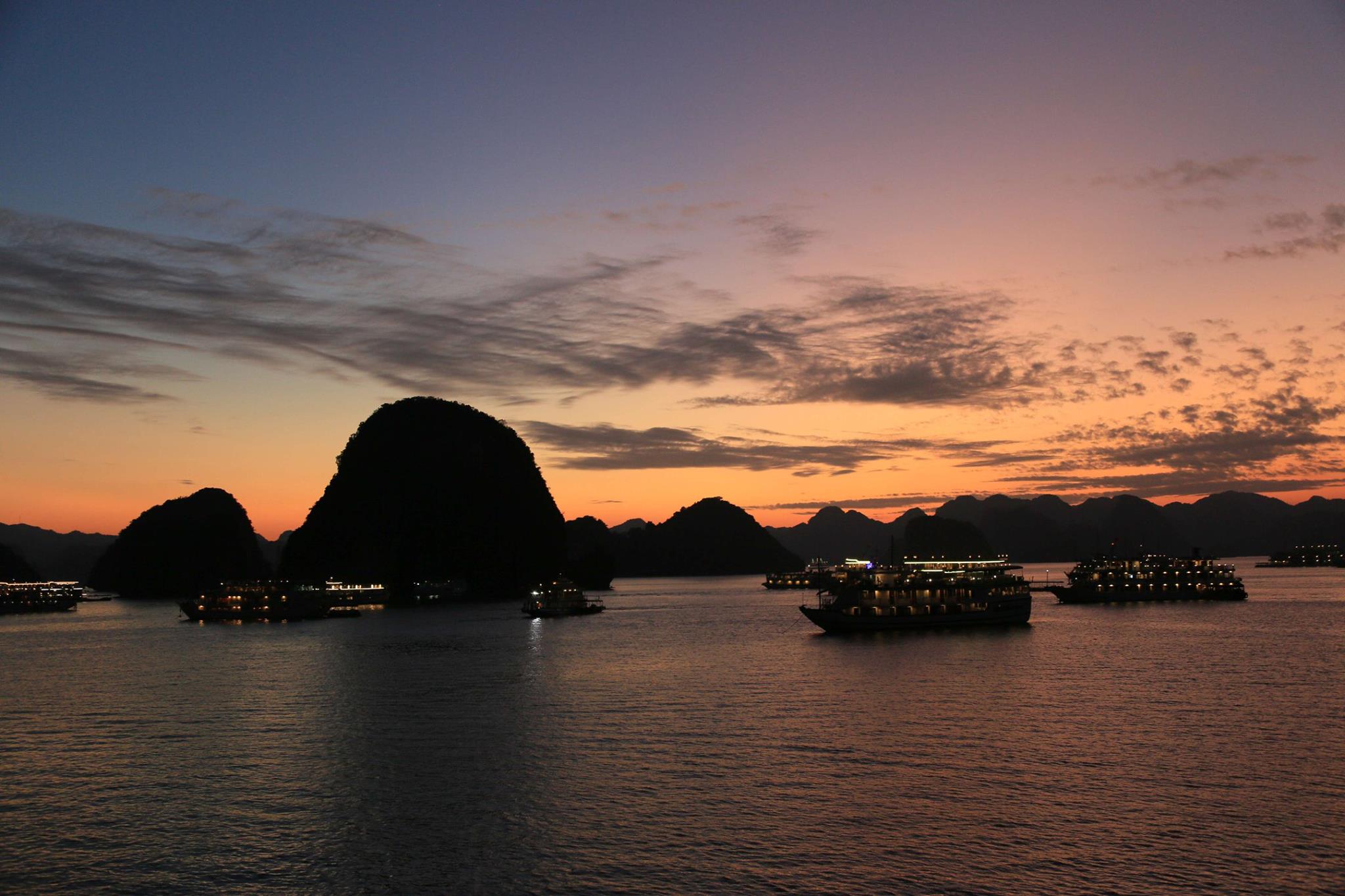 Sunset over calm water with silhouetted limestone islands and anchored boats on the shorelight sky in orange and pink hues