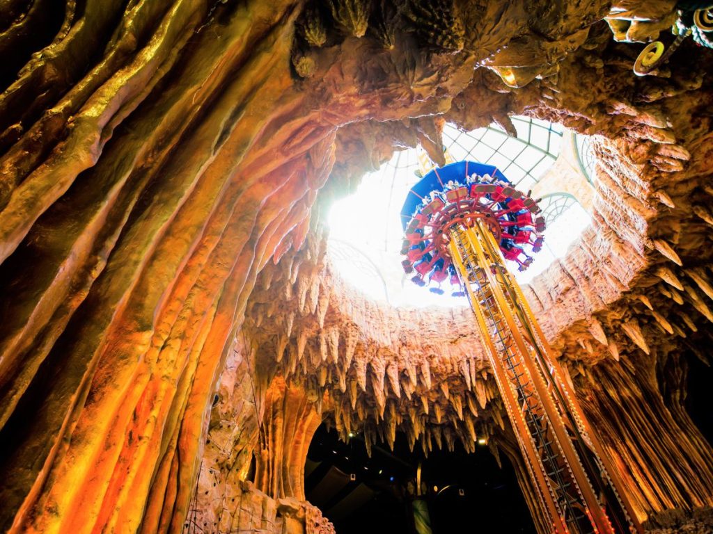 Inside a cavern with stalactites and a tall colorful amusement ride spiraling upward toward a glass skylight. (Alternative: focus on structure and setting)