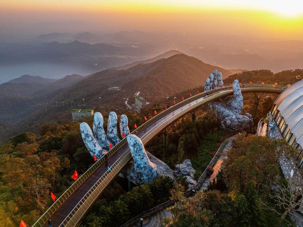 Sunset over a mountain range with a curved skywalk and giant stone hand sculptures along the railing, red flags fluttering nearby.